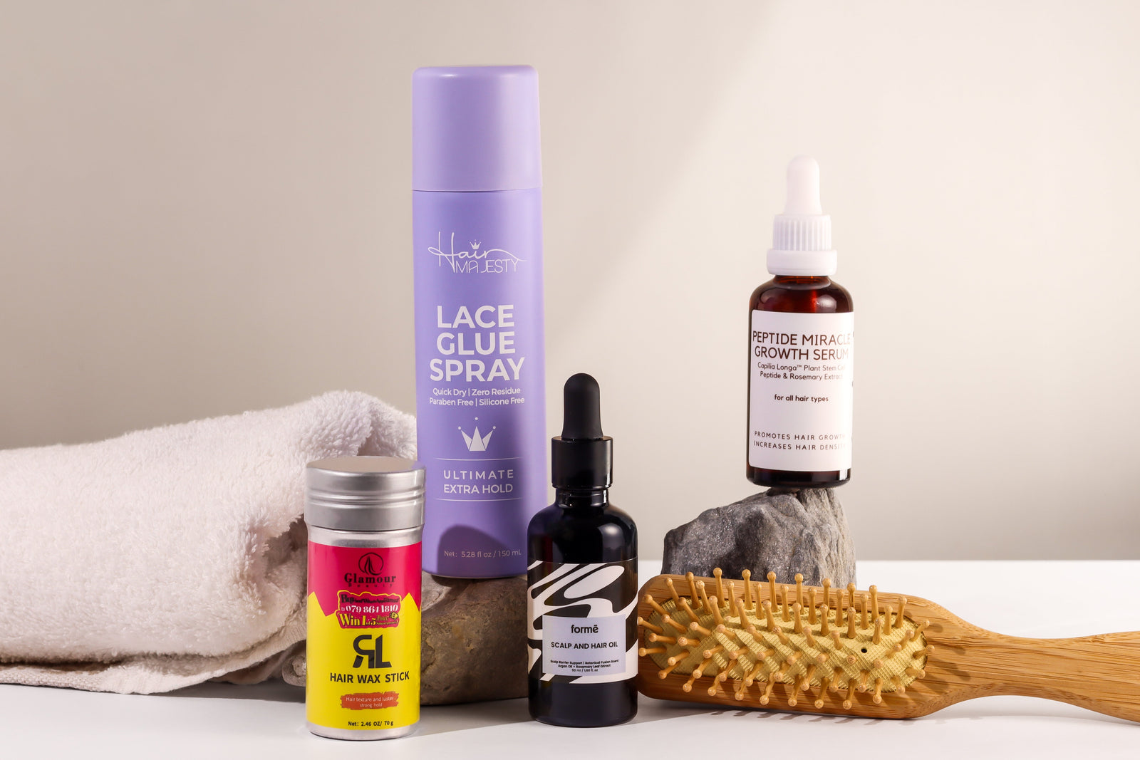 Hair Majesty lace glue spray, Glamour Beauty wax stick, Formē hair and scalp oil, and STANDARD. peptide serum beside a wooden brush.